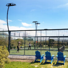 Guests enjoying a sunny outdoor tennis court, surrounded by lush greenery, with blue deck chairs for relaxation.
