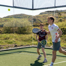 Guests enjoy a lively padel game on scenic outdoor court with lush hills in background.