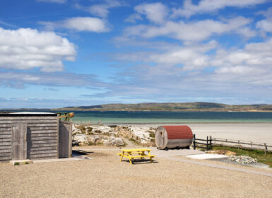 Scenic beach view with picnic spot by hotel sand sauna, overlooking serene sea and rolling hills under blue skies.