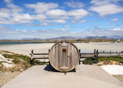 Scenic beach view with unique round sauna, sandy shoreline, and distant mountains; perfect for relaxing holidays.