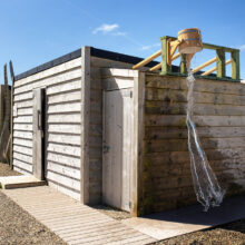 Rustic outdoor wooden shower with water bucket at countryside hotel under clear blue skies.